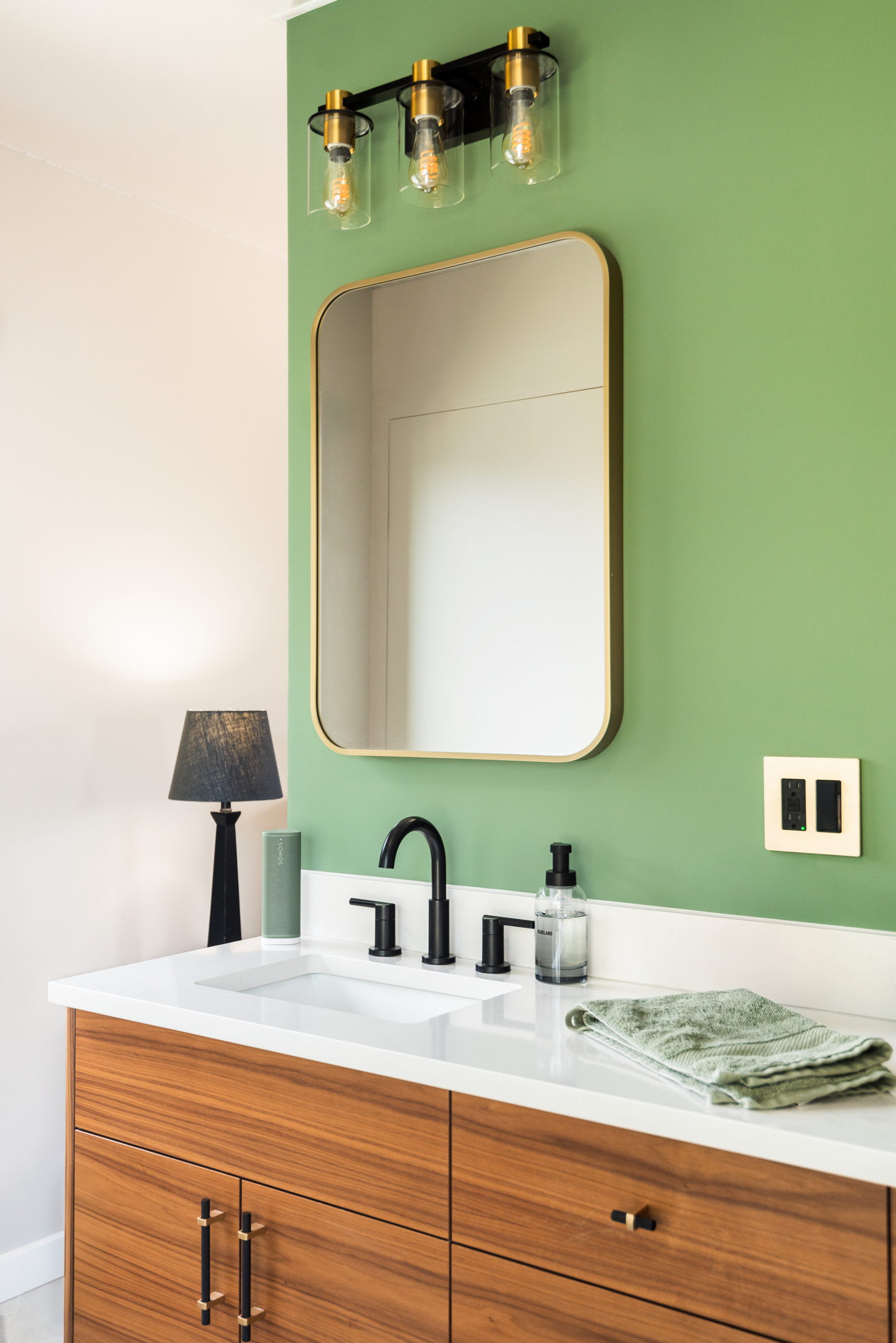 A double walnut vanity against a green wall with brass mirrors and black sconces