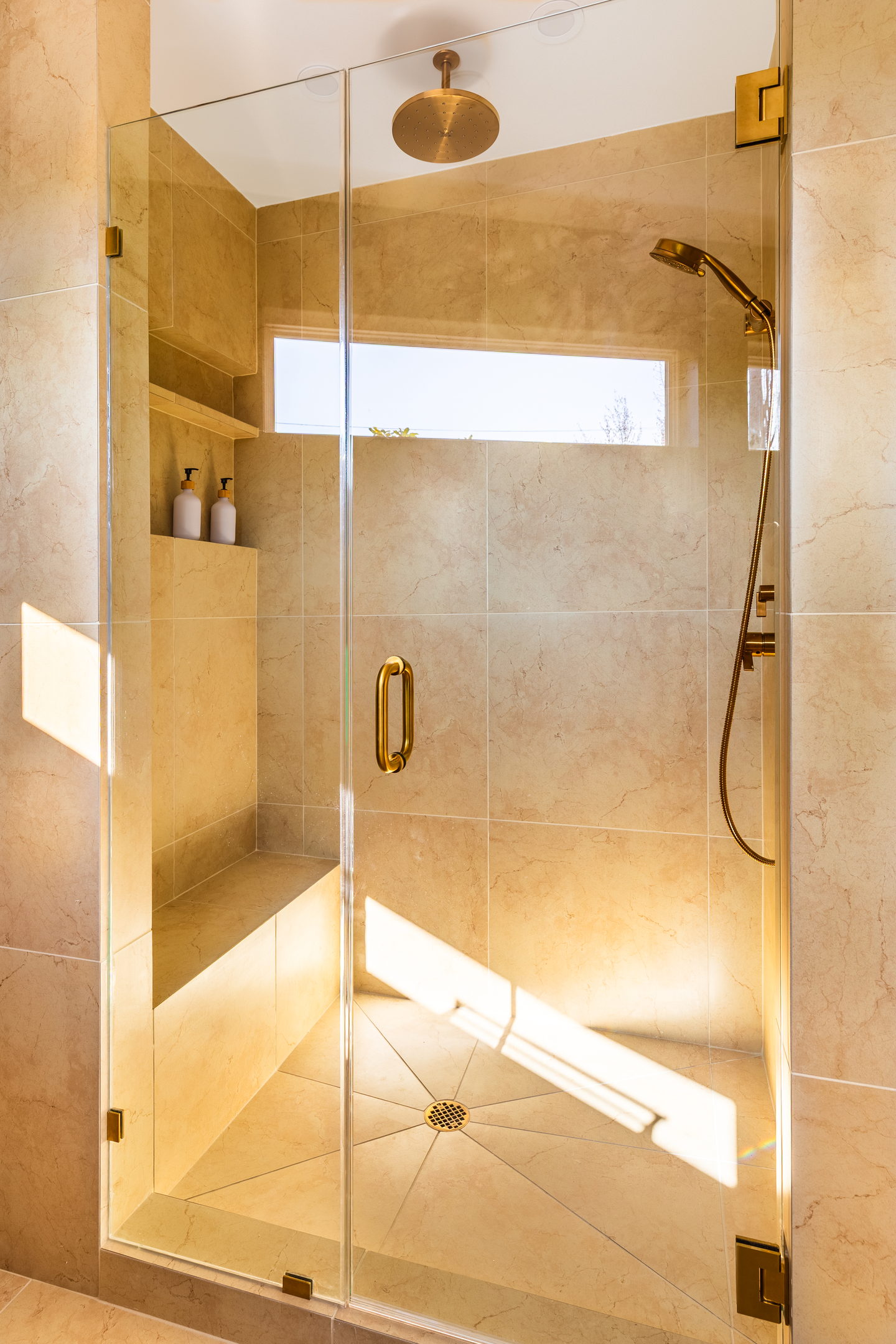 A shower lined in warm stone with a brass rain head and a frameless glass door