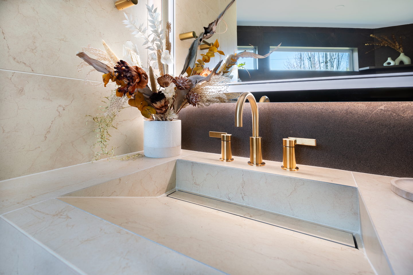 A stone sink with a brass bridge faucet and a dried arrangement