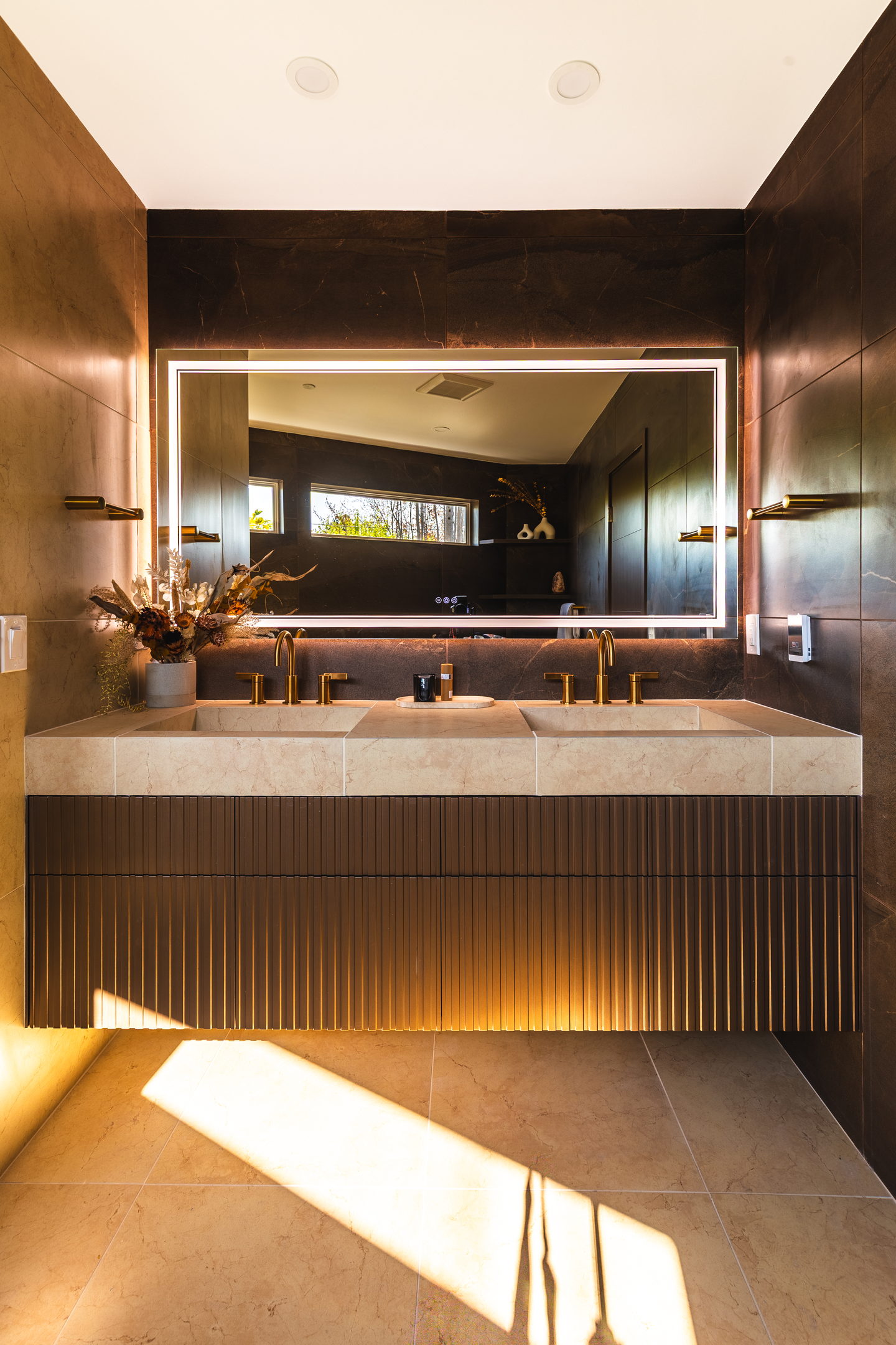 A backlit mirror over a fluted vanity in the dark stone primary bathroom